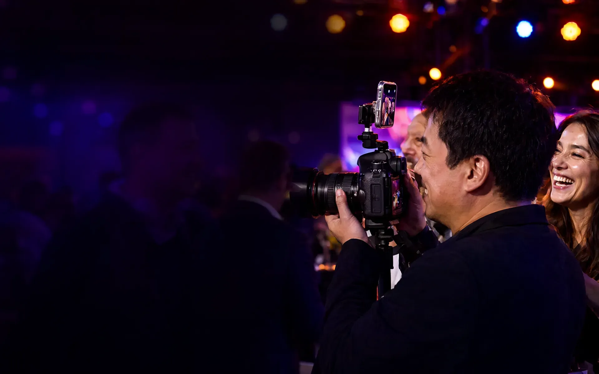 Eventfotograf mit Profikamera und aufgestecktem Smartphone bei einem Abendevent, neben ihm lacht ein Gast