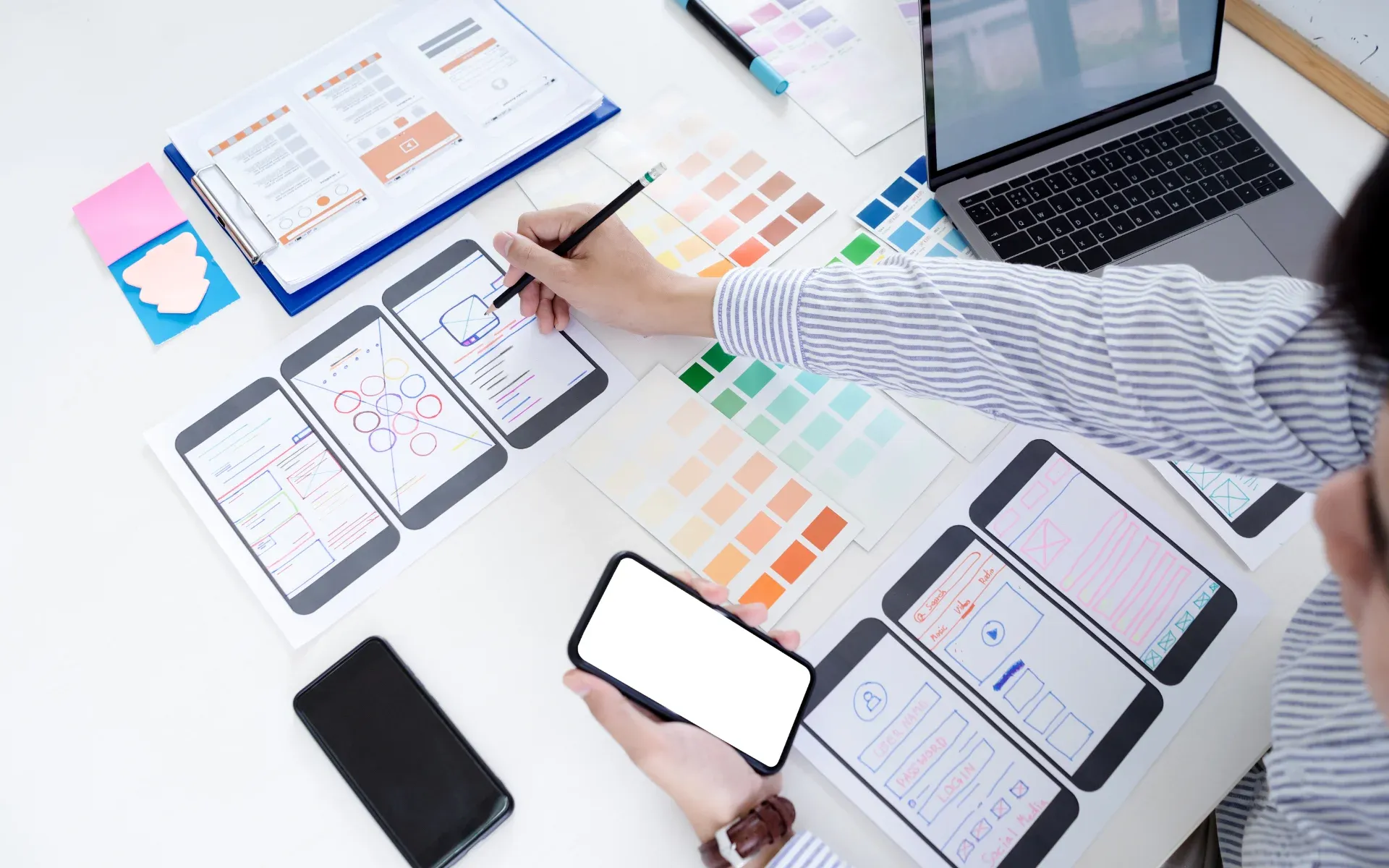UX designer sketching app wireframes on paper next to a smartphone and laptop